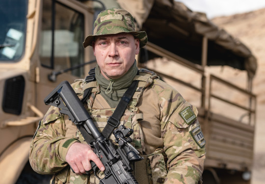 A soldier stands in uniform facing the camera holding a weapon with a New Zealand Army truck in the background