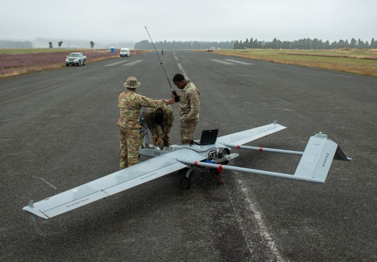 Three soldiers work on the Shadow UAV in the middle of a runway, one soldier is holding a long a device with a long antenna and a laptop sits on the UAV. Two civilian vehicles are parked on the edge of the runway in the background. Low cloud and fog can b