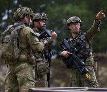 Three soldiers in Army uniform holding weapons having a discussion. The solider on the right points into the distance.