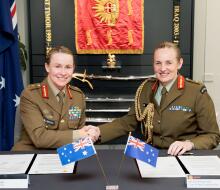 Two women Chiefs of Army for New Zealand and Australia sit at a table in front of two documents and their corresponding countries flag, they are both smiling and shaking hands. 