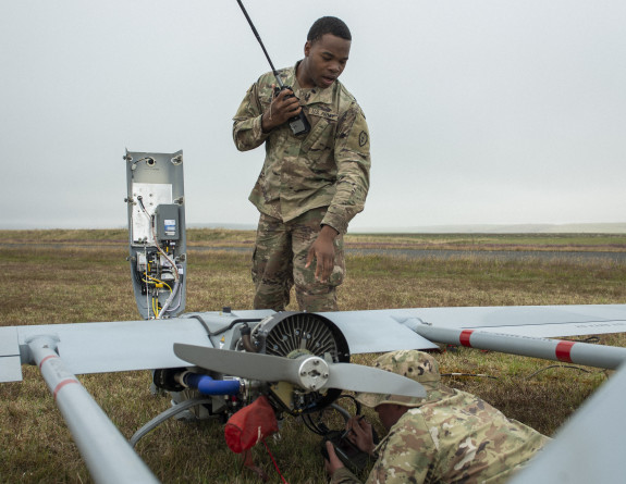 A soldier stands above the propellor of the Shadow UAS while holding a device with a long antenna attached. Another soldier lays on the ground as they work on the a propellor itself. The UAS is large and fully surrounds the solider on the ground. The fiel