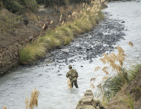 A soldier crosses a river that is set between two hills from a high angle. Another soldier crouches as they descend the hill closer to the camera.