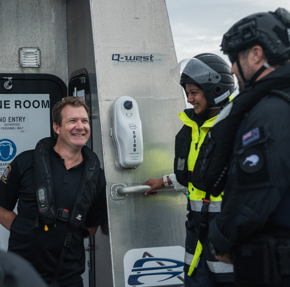 Four people talk aboard Hawk V, the NZ Customs vessel. A man from NZ Customs talks with a person in a high visibility jacked from MPI and members of the Deployable Boarding team, all wearing dark blue Navy uniform, helmets and lifejackets.