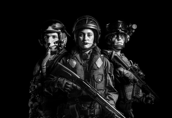 A black and white photo of three NZDF personnel. The left is Air Force and is wearing a flying helmet and mask. In the middle the personnel is Navy and wearing a lifejacket and helmet with a headset. The person on the right is soldier and wearing headgear