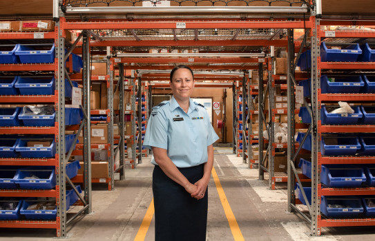 A woman stands in a warehouse with her hands together in front of her, she is wearing a RNZAF uniform. In the background are industrial shelving, nets, trays and boxes.