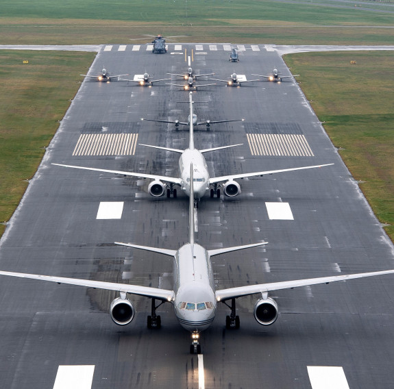 A variety of grey painted Air Force aircraft, large and small are lined up on the centre of a runway. The photograph is taken from the air, showing the aircraft, nose on.