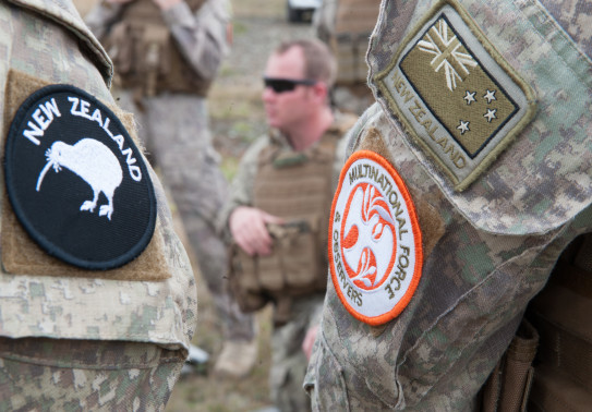On the left an arm (in camo shirt) wears the New Zealand patch with the Kiwi on it and the right arm of a different person has the Multinational Force patch and a New Zealand flag patch. Other NZDF personnel are shown in the background