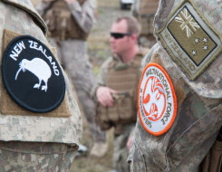 On the left an arm (in camo shirt) wears the New Zealand patch with the Kiwi on it and the right arm of a different person has the Multinational Force patch and a New Zealand flag patch. Other NZDF personnel are shown in the background
