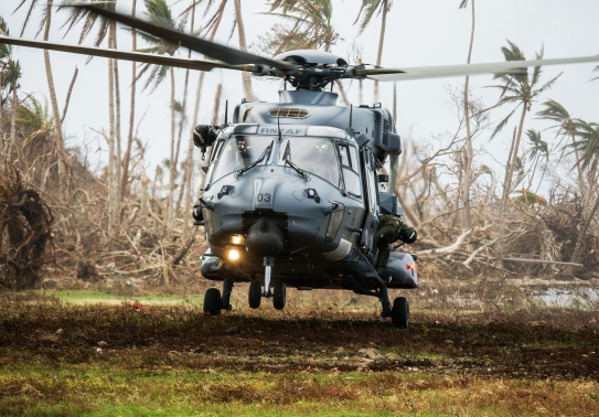 A Royal New Zealand Air Force NH90 helicopter lands on the grass in Fiji, palm trees blowing the background as well as mess from Cyclone Winston