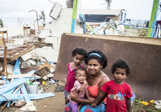 Three children and a mother pose for a photo with the damage post Cyclone Winston in the background. Further in the background you can see the beach and ocean