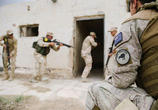 A New Zealand Army soldier sits to the right of the image and watches other four uniformed soldiers enter a building with weapons.