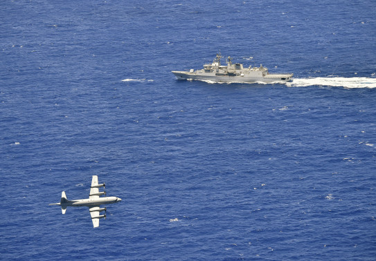A Royal New Zealand Air Force P-3K2 Orion flies over the blue ocean in the bottom left side of the photo with HMNZS Te Kaha during a firing exercise 