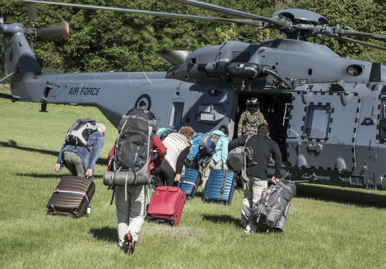People with luggage and other bags walk toward a Royal New Zealand Air Force NH90 helicopter and Helicopter Loadmaster on the grass in Kaikōura after the earthquake