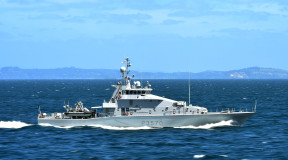 Royal New Zealand Navy's HMNZS Taupō sailing in the ocean on a nice day, blue sky in the background and some hills. 