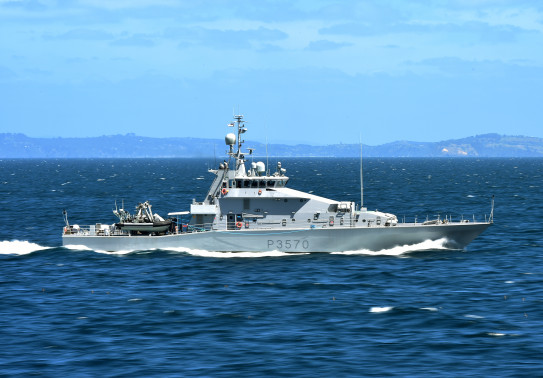 Royal New Zealand Navy's HMNZS Taupō sailing in the ocean on a nice day, blue sky in the background and some hills. 