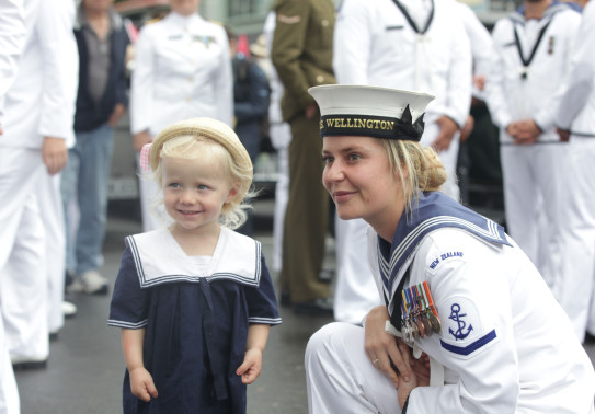 A sailor bends down to engage with a young child who is dressed in a sailor outfit. They are both smiling. You can also see other sailors standing in the background of the photo