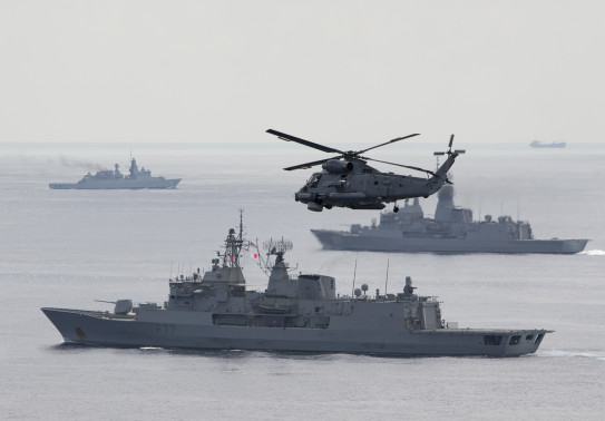 A SH-2G(I) Seasprite Helicopter flies in the foreground of the image with HMNZS Te Kaha. In the background there are two other warships from other countries