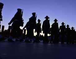 NZDF personnel in uniform march in formation during an early morning ceremony. The personnel are silhouetted and the sky looks dark blue in the background. 