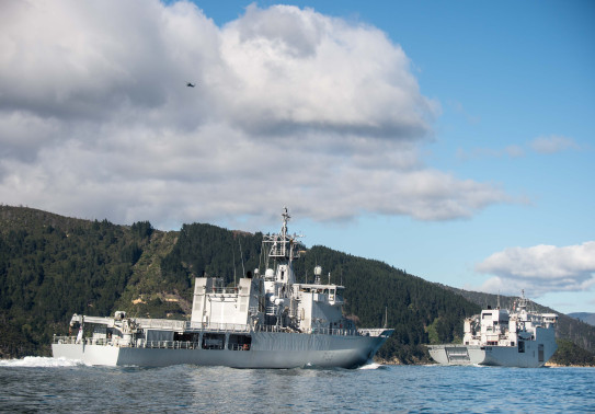 HMNZS Wellington transits behind HMNZS Canterbury in the Marlborough Sounds on a nice day - clouds and blue sky. Green hill area in the background the photo. 