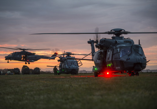 Three Royal New Zealand Air Force NH90 helicopters at dusk. One aircraft in the background is in flight, the other two are on the ground. The sky is an orange morning sunrise.