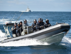 A Royal New Zealand Navy Rigid-Hulled Inflatable Boat (RHIB) full with sailors wearing helmets moves through the ocean with a ship in the background on a nice day, blue sky.