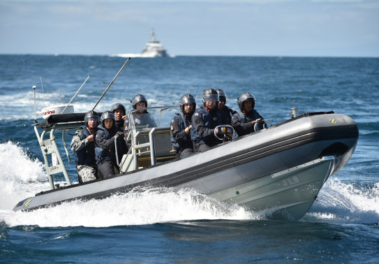 A Royal New Zealand Navy Rigid-Hulled Inflatable Boat (RHIB) full with sailors wearing helmets moves through the ocean with a ship in the background on a nice day, blue sky.