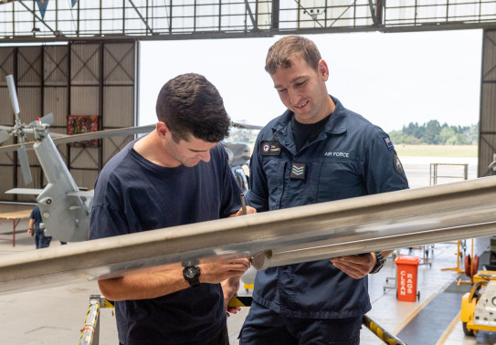 Two Royal New Zealand Air Force avionic technicians look at a piece of equipment in an aircraft hanger. There is a helicopter in the background and a door opening to outside. 