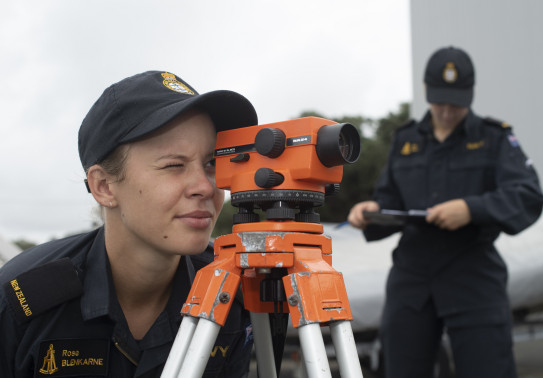 A Royal New Zealand Navy hydrographers being assessed by another Navy sailor in the background. The sailor is looking through a piece of equipment that is orange, they have one eye looking through the scope and the other closed.  