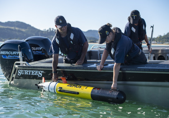 Three Royal New Zealand Navy personnel in a boat on a nice day. Two sailors lean over the boat to reach for a piece of equipment. In the background there are tree covered hills. 