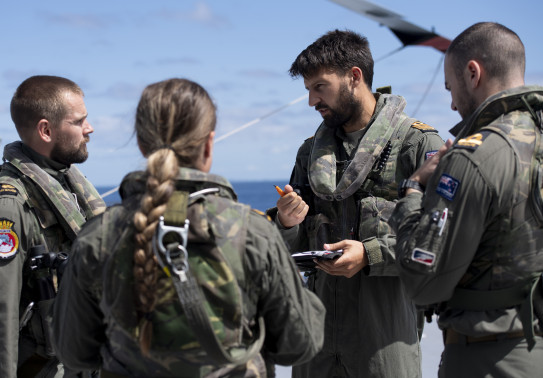 Four Royal New Zealand Navy aircraft personnel talking in a group. The person whose face is shown is holding a pen and a notepad. 