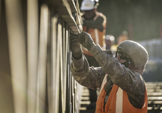 A New Zealand Army soldier works on a bridge with a helmet, glasses and an orange high vis vest. There are other people in the background working on the bridge as well.