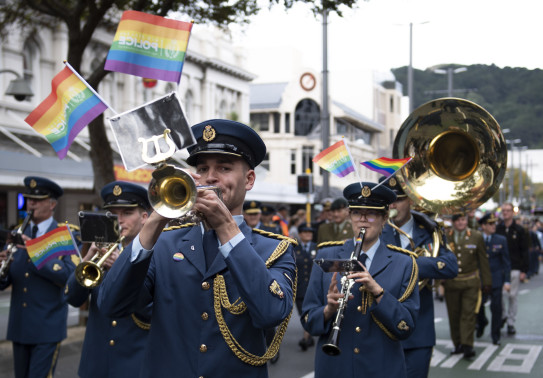 Royal New Zealand Air Force Band members walk down a street playing their instruments with rainbow flags. 