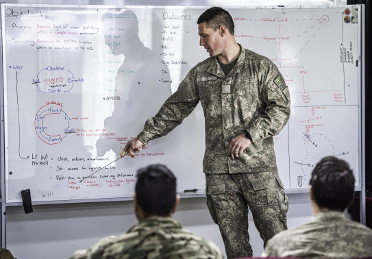 A New Zealand Army soldier points a whiteboard with content on it. In the foreground, blurred are other uniformed people watching on. 