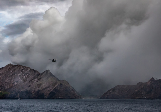 A SH-2G(I) Seasprite Helicopter flies in the air near the Whaakari/White Island. In the image you can see smoke erupting from the volcano
