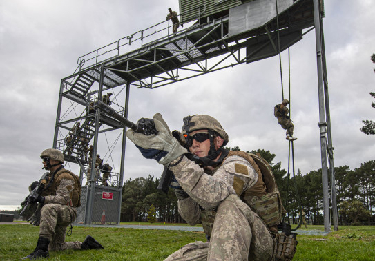 New Zealand Army soldiers fast roping from a tower in the background and in the foreground two soldiers provide ground cover below the tower. One solider on the tower watches as and accesses the soldiers..