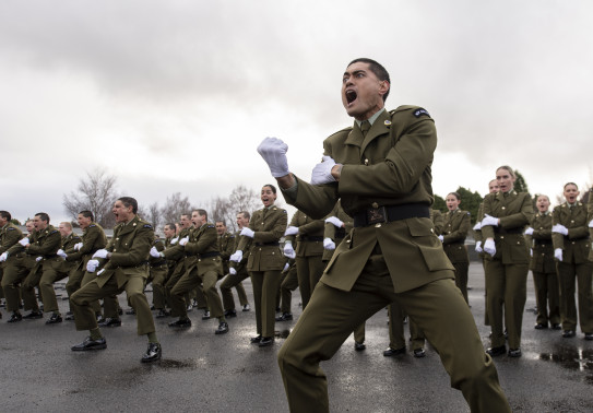 New Zealand Army soldiers perform a haka. The soldier in the foreground is making a fist as part of the haka and the photo is caught while yelling. Other soldiers are seen in the background.