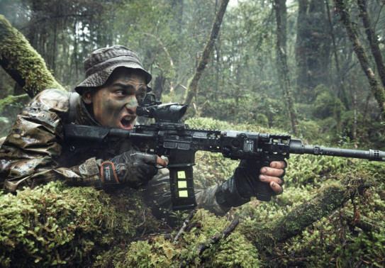 A New Zealand Army soldier in the New Zealand bush with MARS-L weapon. The area is all wet and very green. The soldier has his mouth open as he yells a command. 