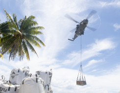 A Royal New Zealand Air Force NH90 helicopter carries an underslung load on the right of the frame. On the left of the frame there are palm trees and other loads.