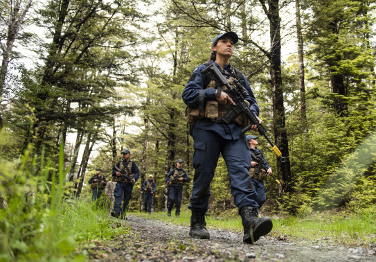 A Royal New Zealand Air Force airman walks with a weapon on a shingled path with other airmen in the background. 