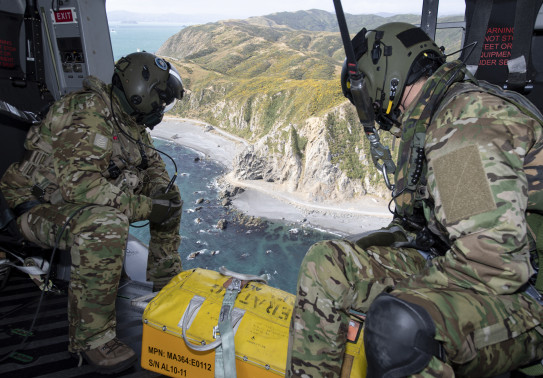 Royal New Zealand Air Force helicopter loadmasters look outside the aircraft during a search and rescue mission. In the photo you can see the rigid coastline and ocean. There is also a box at their feet they would use if they found the missing people