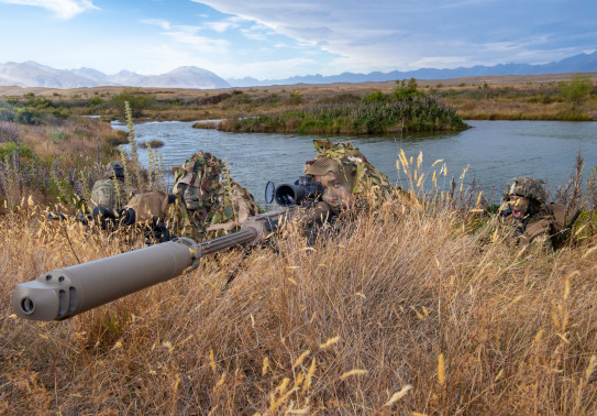 New Zealand Army soldiers lie in the grass around a lake area with the M107A1 Anti-Materiel Rifle. The backdrop shows the mountainous ranges around Tekapo Military Training Area. The grass is dry and its a sunny day with some cloud. 