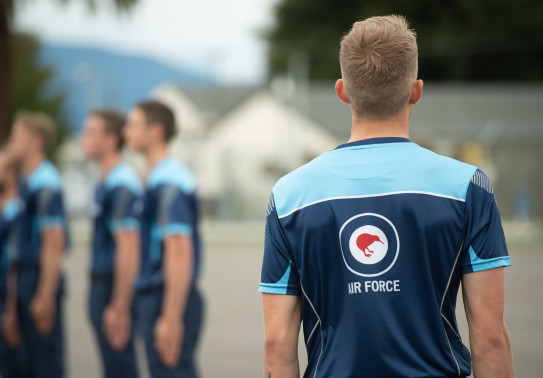 Royal New Zealand Air Force personnel. The photo shows the back of one of them with the Air Force roundel logo. In the background, out of focus there are more Air Force personnel.