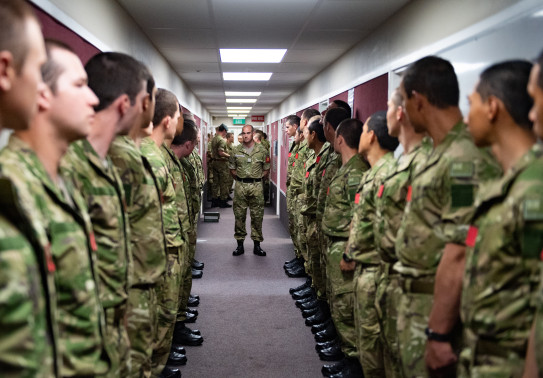 New Zealand Army soldiers stand in a corridor a row of people on each side. One solider stands in the middle of the corridor. 