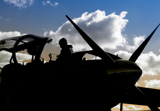 A Royal New Zealand Air Force pilot about to get in a T-6C Texan II aircraft. The pilot and the aircraft is silhouetted in the image. The sky is blue with a bit of cloud. 