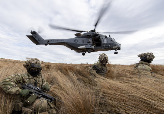 New Zealand Army soldiers sit in the tussock grass within the Waiouru Military Training Area after being dropped off by an NH90 helicopter (seen behind them flying away). 