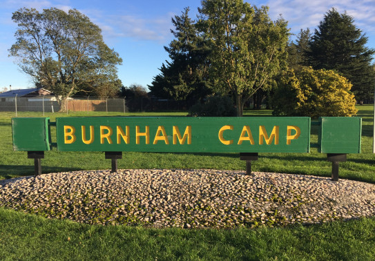 A green and yellow sign that says 'Burnham Camp' is fixed in the ground. In the background you see trees and blue sky