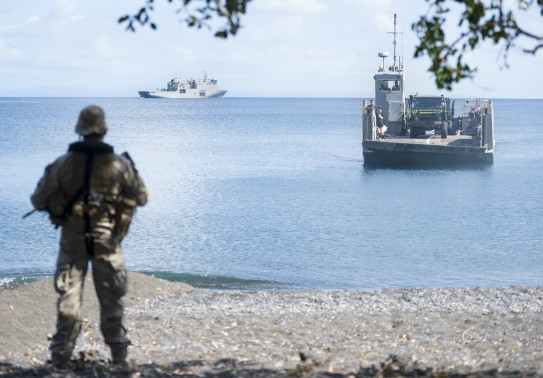 A New Zealand Army soldier watches from the shore as a Royal New Zealand Navy Landing Craft Mechanised (LCM) is sent from HMNZS Canterbury. The image is framed with branches from a tree. 