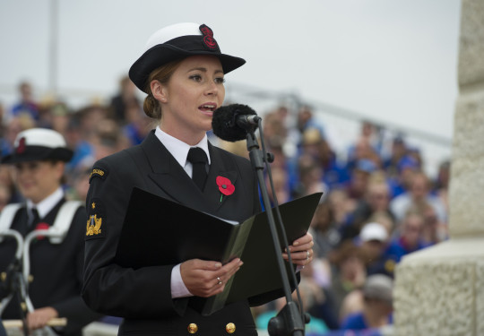 Able Musician Rebecca Nelson singing at a commemoration holding a folder with a microphone in front. in the background, out of focus you can see grounds sitting in stands and another Navy Band member. 
