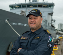 Sailor in working dress standing in front of an Offshore Patrol Vessel while ashore.