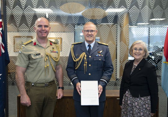 Three people stand in front of a glass wall with patterns on it. The New Zealand flag to the left. Chief of the New Zealand Defence Force Air Marshal Kevin Short (centre) was presented the commendation by Defence Adviser Colonel Neil Peake (left) and High